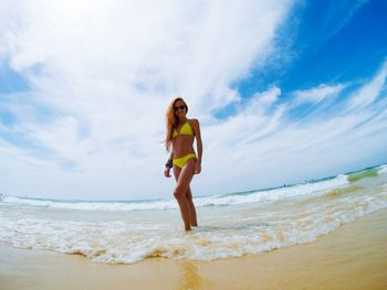 Low angle view of mid adult woman in bikini standing at beach against cloudy sky