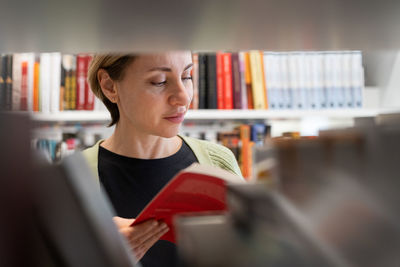 Female mature student looking for book at library bookshelf studying at university campus or college
