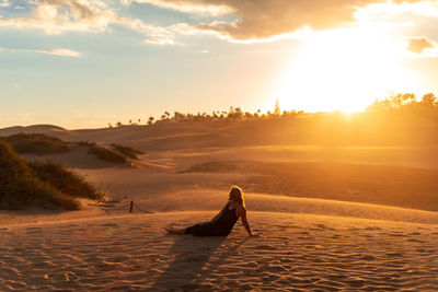 Full length of woman sitting on sand against sky during sunset