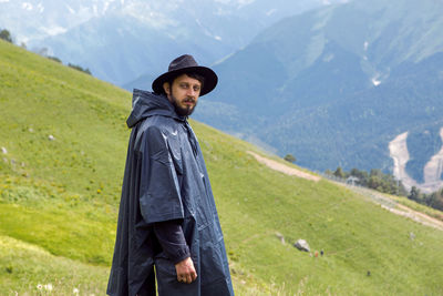 Man with a beard shepherd standing in the mountains in a black raincoat in the rain