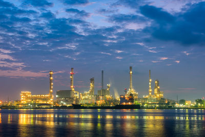 Illuminated factory by buildings against sky at night