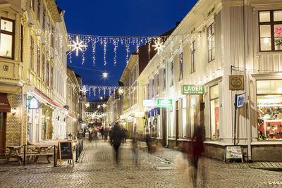 Blurred motion of people walking on illuminated city street at dusk