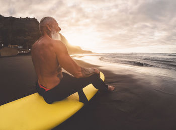 Senior man sitting on beach against sky during sunset