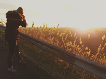 Full length of woman standing by railing while photographing during sunset