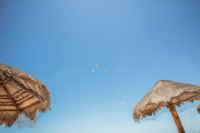 Low angle view of thatched roofs and birds flying against blue sky
