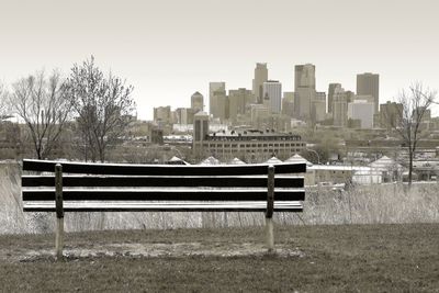 Park bench by buildings in city against clear sky