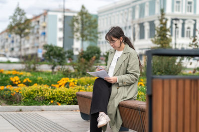 A girl sits on a bench on the street with documents in her hands and using her phone