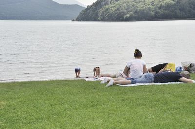 Rear view of friends sitting on grass by lake against sky