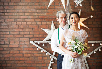 Portrait of a smiling young woman holding bouquet against brick wall