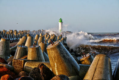 Lighthouse and waves splashing on groynes by sea against clear sky