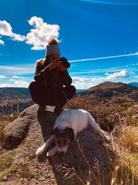Young woman with dog photographing while crouching on rock against sky