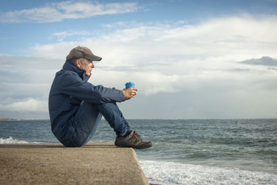 Side view of man sitting at beach against sky during sunset