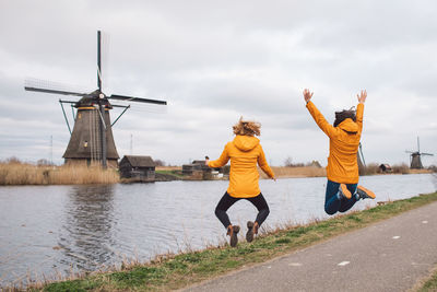 Rear view of women jumping on riverbank against sky and traditional windmills 