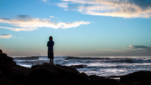 Rear view of man standing on rocks by sea against sky