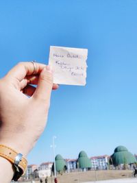 Cropped image of hand holding paper against clear blue sky