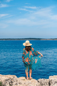 Rear view of woman wearing hat standing rock against sea