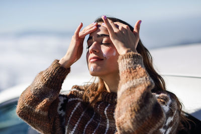 Young woman with eyes closed applying moisturizer while standing outdoors during winter
