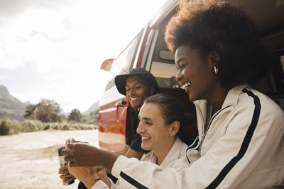 Happy male and female multiracial friends sharing smart phone while sitting in van