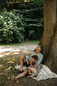 Grandfather reading story book to grandson while sitting under tree at park