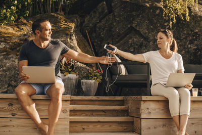 Mature woman giving smart phone to man with laptop while sitting on patio