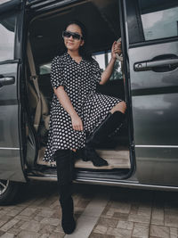 A girl wearing a polkadot dress, sun glasses and high boots sitting in a car 