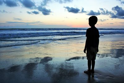 Rear view of silhouette boy standing at beach against sky during sunset