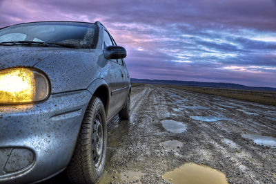 Dirty car on muddy road against cloudy sky during sunrise