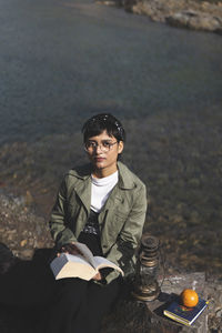 Portrait of young woman reading book while sitting by lake