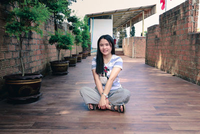 Portrait of smiling woman sitting on hardwood floor by potted plants outdoors