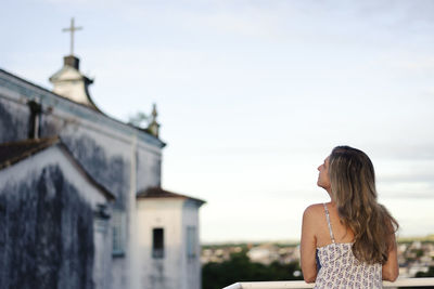 A woman on the porch with her back to the camera against the sky and church in the background. 