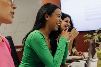 Happy female business professional leaning on elbows while sitting with colleagues during meeting at office