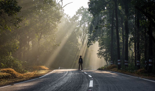 Boy standing on road against trees during sunny day