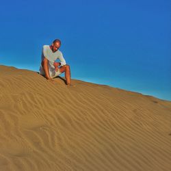 Low angle view of tired man sitting on sand dune against clear blue sky