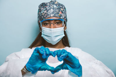 Portrait of young woman wearing mask making heart shape against colored background
