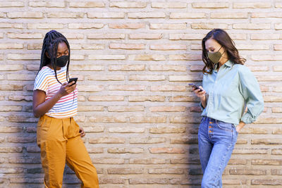 Friends wearing mask using smart phone standing by wall outdoors