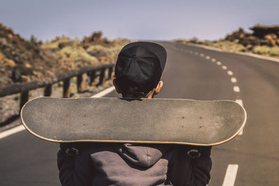 Rear view of boy holding skateboard while standing on road