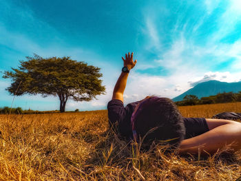 Man gesturing while lying on grassy field against blue sky