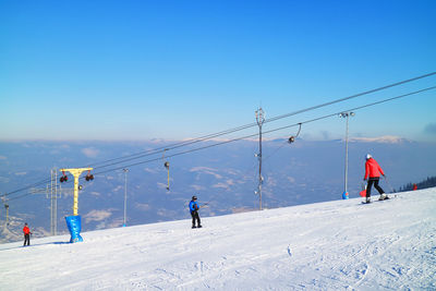 People on surface lift at snow covered field against clear blue sky