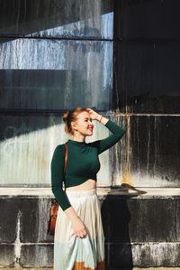 Smiling young woman standing by wall during sunny day