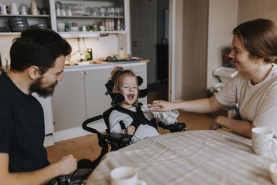 Smiling parents with happy disabled child in wheelchair sitting at kitchen table