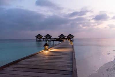 Pier towards stilt houses amidst sea against cloudy sky