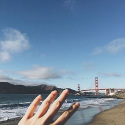 Cropped image of hand against suspension bridge over sea