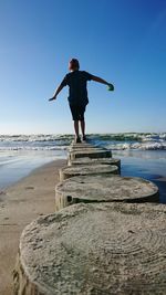 Rear view of boy balancing on tree stumps at beach against blue sky