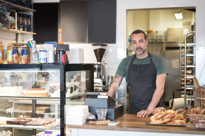 Portrait of confident mature male owner standing at checkout in bakery