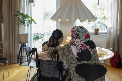 Rear view of mother sitting with daughter having paraplegia at home