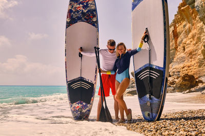 Young active couple stands on a beach hugging and holding stand up paddle boards