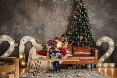 Mother and son with gift sitting on chair against christmas tree