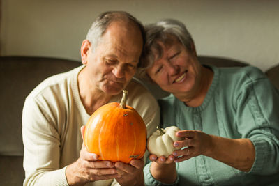 Smiling senior couple holding two pumpkins side by side, preparing decor to halloween holiday.