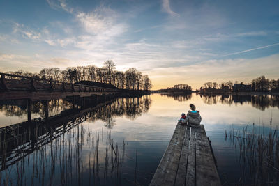 Rear view of mother and son sitting by lake against sky