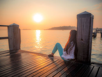 Side view of woman sitting on jetty over sea against sky during sunset